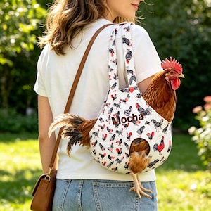 May include: A white fabric chicken carrier bag featuring a repeating pattern of roosters and hens in black, white, and red. The bag has a circular opening for the chicken's legs and the name Mocha embroidered in brown thread on the front side.