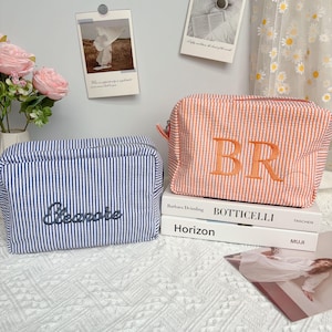 Può includere: Due trousse rettangolari. Una è a righe blu e bianche con il nome "Eleanore" ricamato in corsivo. L'altra è a righe arancioni e bianche con le lettere "BR" ricamate. Entrambe hanno una chiusura con cerniera.