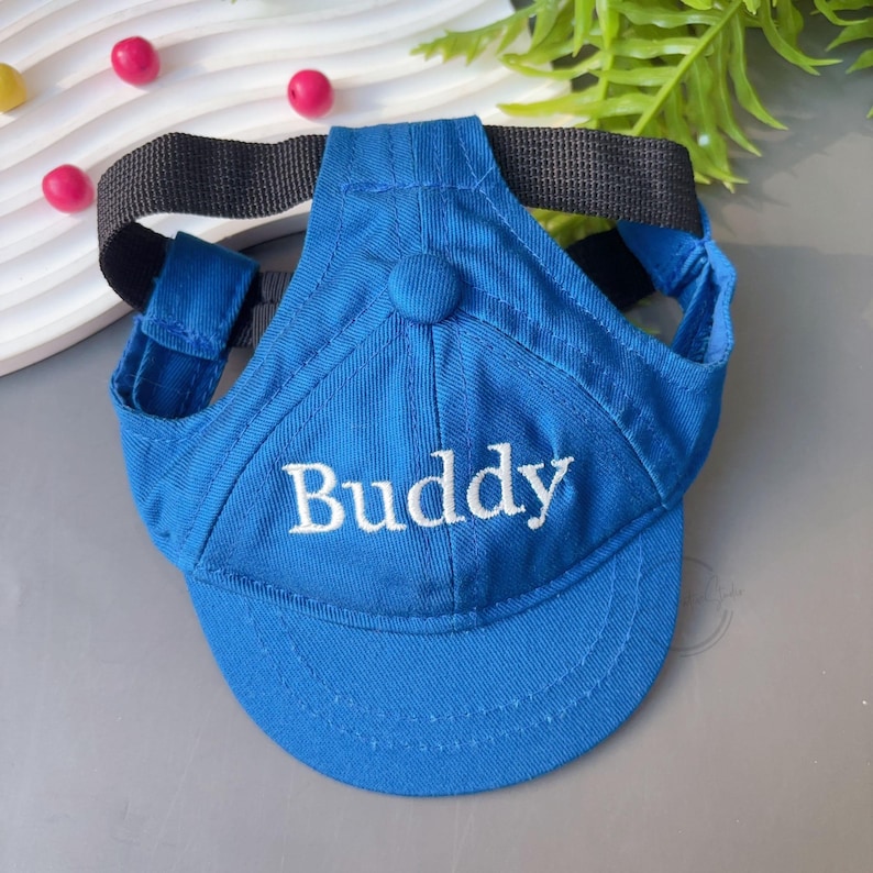 Peut inclure: Casquette de baseball bleue pour chien avec le mot "Buddy" brod&eacute; en blanc. La casquette a une sangle noire et des trous pour les oreilles. Un bouton est sur le dessus.