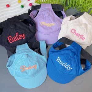 以下が含まれることがあります： 黒、ラベンダー、白、水色、青など、さまざまな色の野球帽スタイルの犬用ハーネスのコレクション。 各キャップには、異なる名前が刺繍されています。Bailey、Maggie、Charlie、Daisy、Buddy。