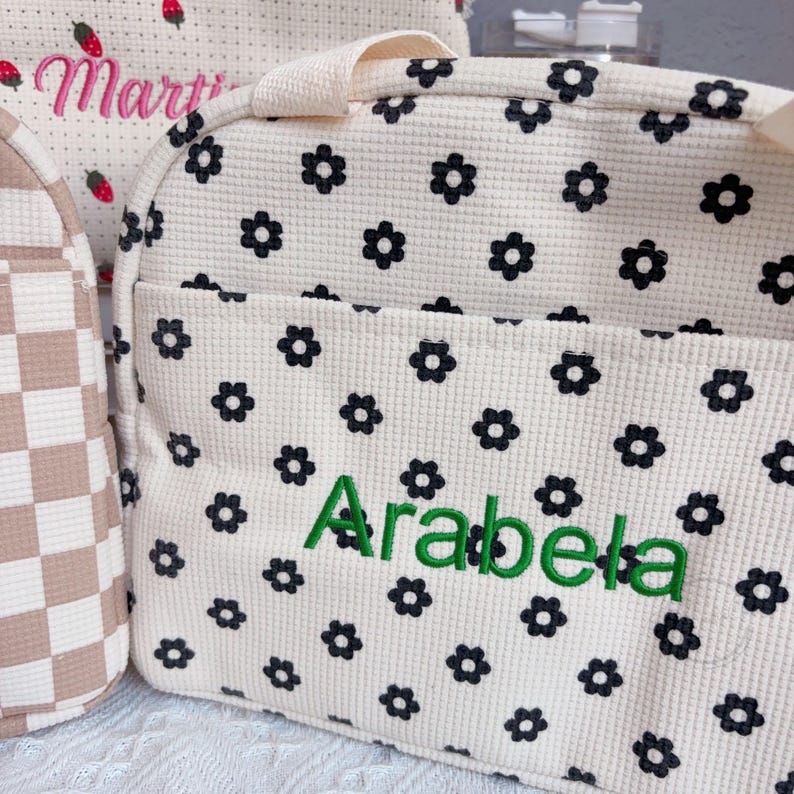 May include: Cream-colored tote bag with a black floral pattern and the name "Arabela" embroidered in green. The bag has a textured waffle weave and a handle. Another bag with the name "Marti" is visible in the background.