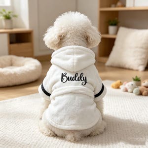 May include: A white plush pet bathrobe with a hood and a tie belt. The back of the robe features the name Buddy embroidered in black script lettering. The sleeves are finished with black trim, and the fabric has a soft, textured appearance.
