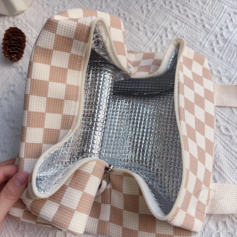 May include: Open insulated lunch bag with a beige and white checkered pattern. The interior is lined with a silver, reflective material. The bag has a zipper closure and a beige handle.