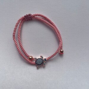 Pode incluir: Pulseira de corda rosa com um pequeno pingente de tartaruga com detalhes azuis e pretos. A pulseira tem contas de metal cor de ouro rosa e um fecho de nó deslizante. Um acessório delicado e elegante.