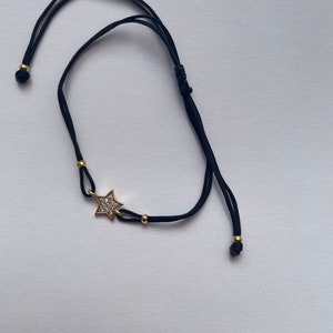 May include: Black adjustable bracelet featuring a gold star pendant encrusted with small, clear crystals. The bracelet has gold-colored beads and adjustable sliding knots for a comfortable fit. A delicate piece of jewelry.