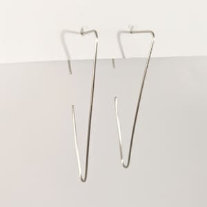Puede incluir: Un par de pendientes plateados minimalistas. Cada pendiente tiene un diseño delgado en forma de V con una base curva. Los pendientes están suspendidos de pequeños postes rectos, creando una estética moderna y geométrica. Los pendientes están sobre un fondo blanco.