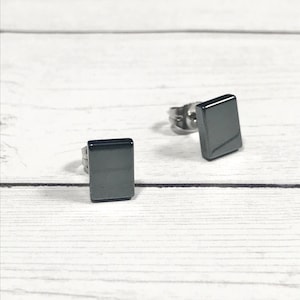 May include: Two square, dark gray stud earrings. The earrings are set against a white wooden background. The earrings have a simple, minimalist design.