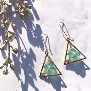 May include: A pair of handmade earrings featuring a triangular gold frame with a light green, textured stone. The earrings have a simple hook design and are displayed on a white marble surface.