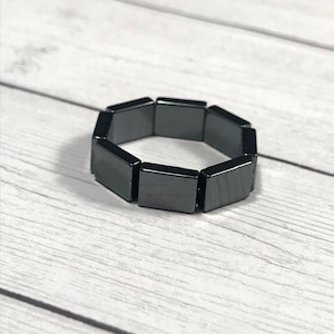 May include: A black bracelet composed of rectangular, polished segments. The bracelet is displayed on a light-colored, wood-grain surface. The segments are connected to form a flexible, circular shape.