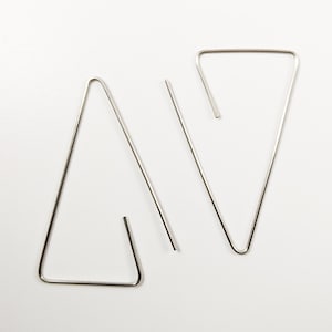 Puede incluir: Un par de pendientes triangulares plateados minimalistas. Los pendientes están hechos de alambre de metal fino y doblado, formando formas geométricas abiertas. El diseño es simple y moderno.