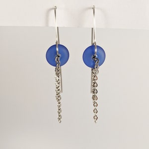 Peut inclure: Paire de boucles d'oreilles pendantes avec une perle ronde bleu mat et une chaîne argentée. Les boucles d'oreilles ont un design simple et élégant avec une fermeture à crochet. Fond blanc.