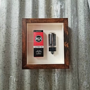 May include: A vintage RCA Radiotron electron tube in a brown wooden frame. The tube is clear glass with a black base and a metal grid inside. The box is red with white text and the RCA logo.