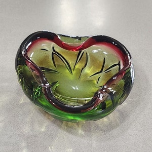 May include: A green and red glass ashtray with a swirl design. The ashtray is shaped like a heart and has a deep well.