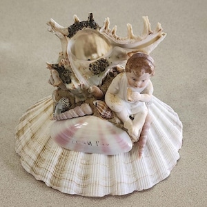 Puede incluir: Una figurita de porcelana de un niño sentado en una concha grande con conchas más pequeñas y arena alrededor.