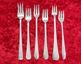 May include: Set of seven silver-toned forks with varying designs. The forks are arranged in a row on a red velvet surface.