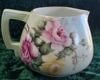 May include: A white porcelain pitcher with a gold rim and a floral design featuring pink and yellow roses. The pitcher has a handle and a rounded body.