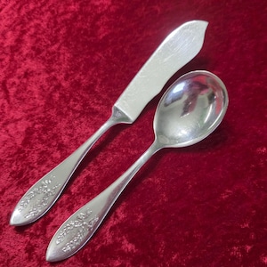 Puede incluir: Un juego de cuchillo de mantequilla y cuchara de plata con detalles florales en los mangos. El cuchillo tiene una hoja redondeada y la cuchara tiene un cuenco redondeado. El juego se muestra sobre una superficie de terciopelo rojo.