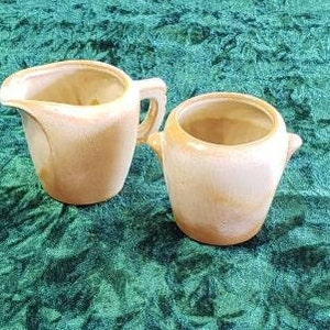 Puede incluir: Un juego vintage de jarra de crema y azucarero de cerámica en un esmalte marrón claro. La jarra tiene un asa y un pico, mientras que el azucarero tiene dos asas pequeñas.