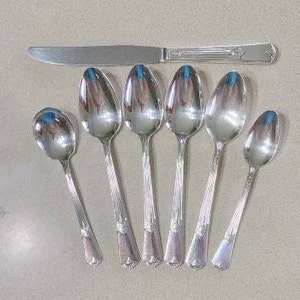 Può includere: Set di sette cucchiai e un coltello argentati. I cucchiai hanno una ciotola arrotondata e un manico intrecciato. Il coltello ha una lama dritta e un manico argentato.