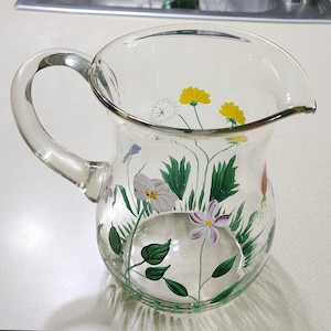 May include: A clear glass pitcher with a handle, painted with a floral design of yellow, white, pink, and blue flowers with green leaves.