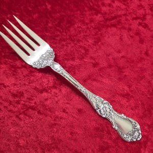 Puede incluir: Un tenedor plateado con cuatro púas, con intrincados detalles florales y de volutas en el mango y las púas. El tenedor se encuentra sobre un fondo de terciopelo rojo, resaltando su elaborado diseño. Este elegante utensilio es adecuado para cenas formales.