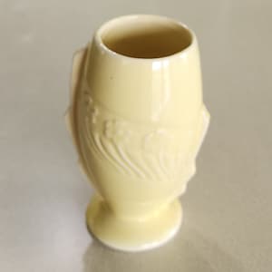 May include: A small, pale yellow ceramic vase with an embossed design. The vase has a rounded body and a flared base.