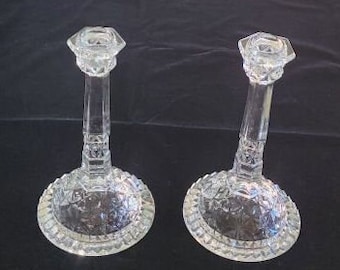 Könnte beinhalten: Zwei klare Glaskerzenhalter mit geschliffenem Glasdesign. Die Kerzenhalter haben einen quadratischen Sockel und eine runde Oberseite.