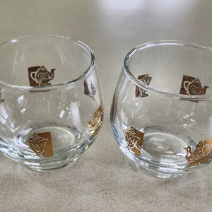 Op de afbeelding: Een set van twee heldere glazen kannetjes voor room en suiker met gouden theepotten en andere theemotieven.
