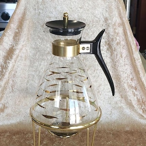 Puede incluir: Una cafetera de vidrio con un diseño dorado y negro sobre un soporte de metal dorado. La cafetera tiene una tapa negra y un asa negra.