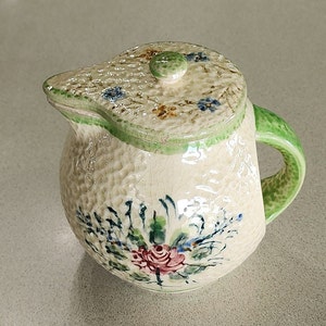 May include: A vintage ceramic teapot with a green lid and handle. The teapot is decorated with a floral pattern in shades of blue, pink, and green.