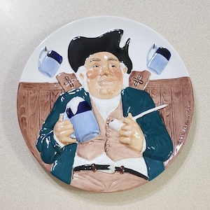 Puede incluir: Plato de cerámica decorativo que representa a un hombre jovial con sombrero negro y abrigo verde, sosteniendo una jarra azul y una pipa. El plato tiene un fondo blanco con detalles marrones y azules, y un diseño de barra de madera.