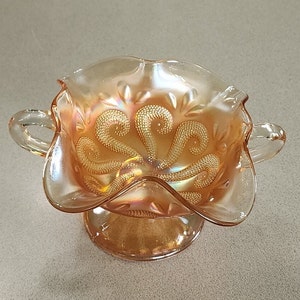 May include: A vintage iridescent glass candy dish with two handles and a footed base. The dish has a swirl design and is in a peach color.