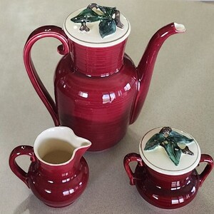 May include: A red ceramic tea set with a teapot, creamer, and sugar bowl. Each piece features a green leaf design on the lid.