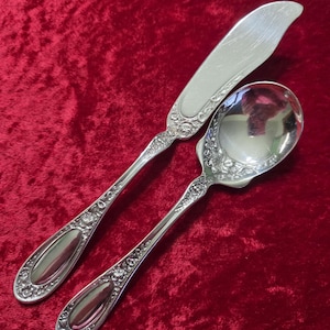 Puede incluir: Juego de cubiertos de plata con un cuchillo de mantequilla y una cuchara, ambos con detalles florales ornamentados en los mangos. La cuchara tiene un cuenco redondeado. El juego se exhibe sobre una superficie de terciopelo rojo.