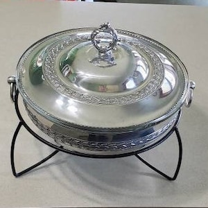 Könnte beinhalten: Ein silberfarbenes, rundes Chafing Dish mit Deckel und dekorativem Rand. Das Gericht steht auf einem schwarzen Metallständer. Der Deckel hat einen Griff und das Gericht hat zwei Seitengriffe.