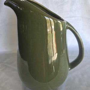 Puede incluir: Jarra de cerámica verde oliva con un pico curvo y un cuerpo redondeado. La jarra tiene un asa grande y un acabado brillante. El diseño es simple y elegante, adecuado para servir bebidas.