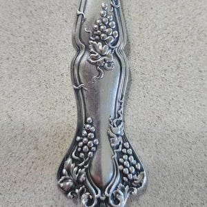 May include: Silverware handle with an ornate design of grapes and vines. The handle is silver and has a polished finish.