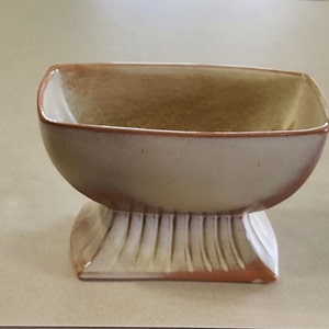 May include: A light brown ceramic planter with a square base and a rounded top. The planter has a textured, ribbed base and a smooth, glazed finish.