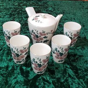 May include: A set of six white ceramic teacups and a teapot with a floral design. The cups and teapot feature a green and pink floral pattern with a white background.
