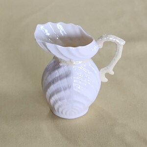 May include: A white porcelain pitcher shaped like a seashell with a detailed, textured surface. The pitcher has a curved handle that resembles a seashell.