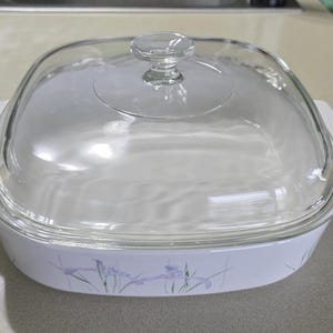 May include: A square, white casserole dish with a clear glass lid. The dish has two white handles and a floral design in shades of purple and blue. The lid has a small, round handle.