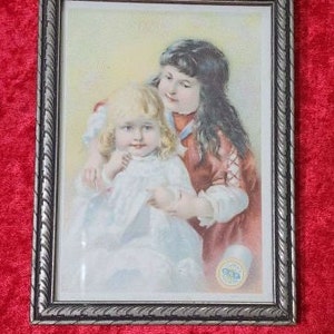 May include: A framed vintage print of two young girls. The girl in the front is wearing a white dress and the girl in the back is wearing a red dress. The print is in a silver frame with a decorative border.