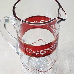May include: A clear glass pitcher with a red and gold band around the top and a decorative design on the side. The pitcher has a handle and is perfect for serving beverages.