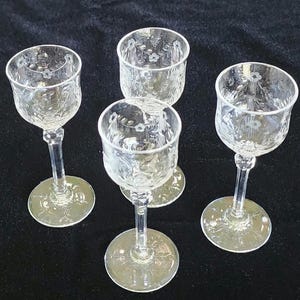 Puede incluir: Cuatro vasos de chupito de vidrio transparente con diseños florales grabados. Los vasos tienen una base con tallo y están dispuestos sobre una superficie negra.