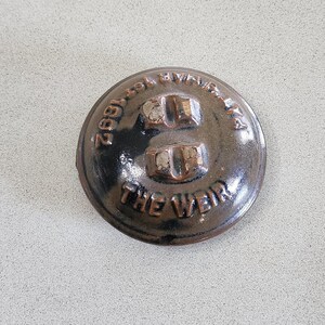 Könnte beinhalten: Ein brauner Keramikdeckel mit dem Text "2892 The Weir" und zwei Metallknöpfen.