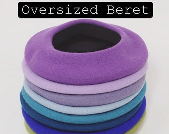 Pode incluir: Uma pilha de oito boinas de tamanho grande em vários tons de roxo, azul e verde. As boinas são feitas de feltro e estão dispostas em ordem de tamanho graduado, com a boina maior no topo.