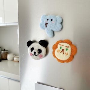 Op de afbeelding: Drie handgemaakte vilten magneten in de vorm van dieren worden op een koelkast getoond. De magneten omvatten een blauwe olifant, een panda en een leeuw. De panda heeft zwarte oren en roze wangen. De leeuw heeft een oranje manen.
