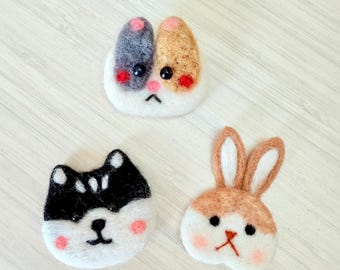 May include: Three felt animal face pins: a cat with calico markings, a black and white husky, and a brown and white bunny. Each pin has a white base, pink cheeks, and black facial features. The pins are handmade.