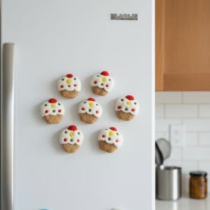 Könnte beinhalten: Sieben cupcake-förmige Magnete mit weißem Zuckerguss, bunten Streuseln und einer roten Kirsche oben sind an einem weißen Kühlschrank befestigt. Die Magnete haben eine braune Basis und sind kreisförmig angeordnet.