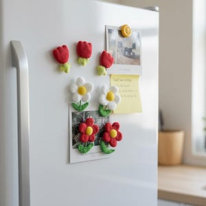 Pode incluir: Uma coleção de ímanes de flores feitos à mão em vermelho, branco e amarelo fixados a um frigorífico branco. Os ímanes incluem tulipas vermelhas, margaridas brancas e margaridas vermelhas com folhas verdes. Um íman amarelo segura uma foto e um post-it.